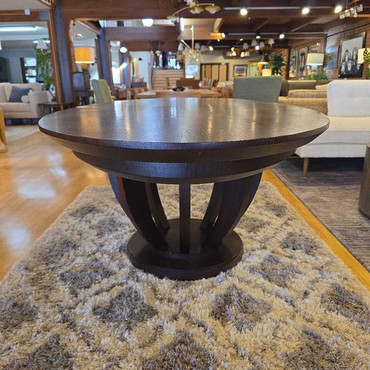 Round wooden table in a showroom setting with furniture and decor in the background