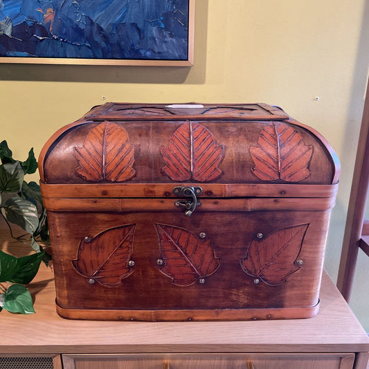 Wooden chest with leaf designs on a wooden surface, with a painting in the background.