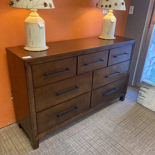 Wooden dresser with five drawers against an orange wall with two lamps on top.