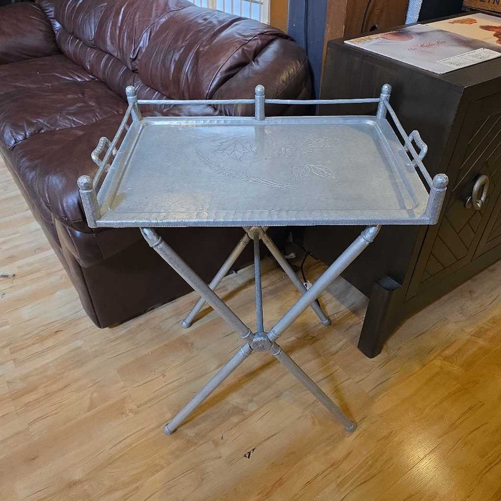Metal tray table on a wooden floor next to a brown leather couch.