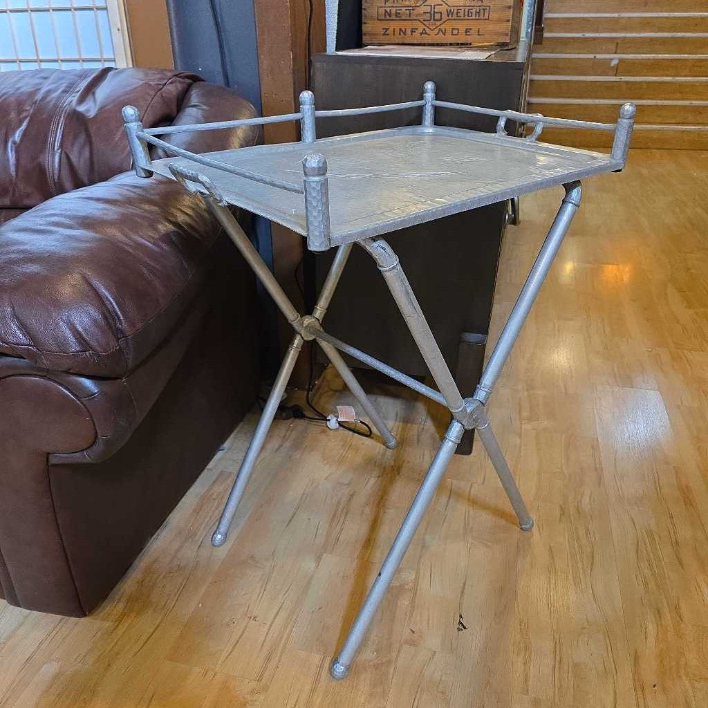Metal folding tray table on a wooden floor next to a brown leather couch.