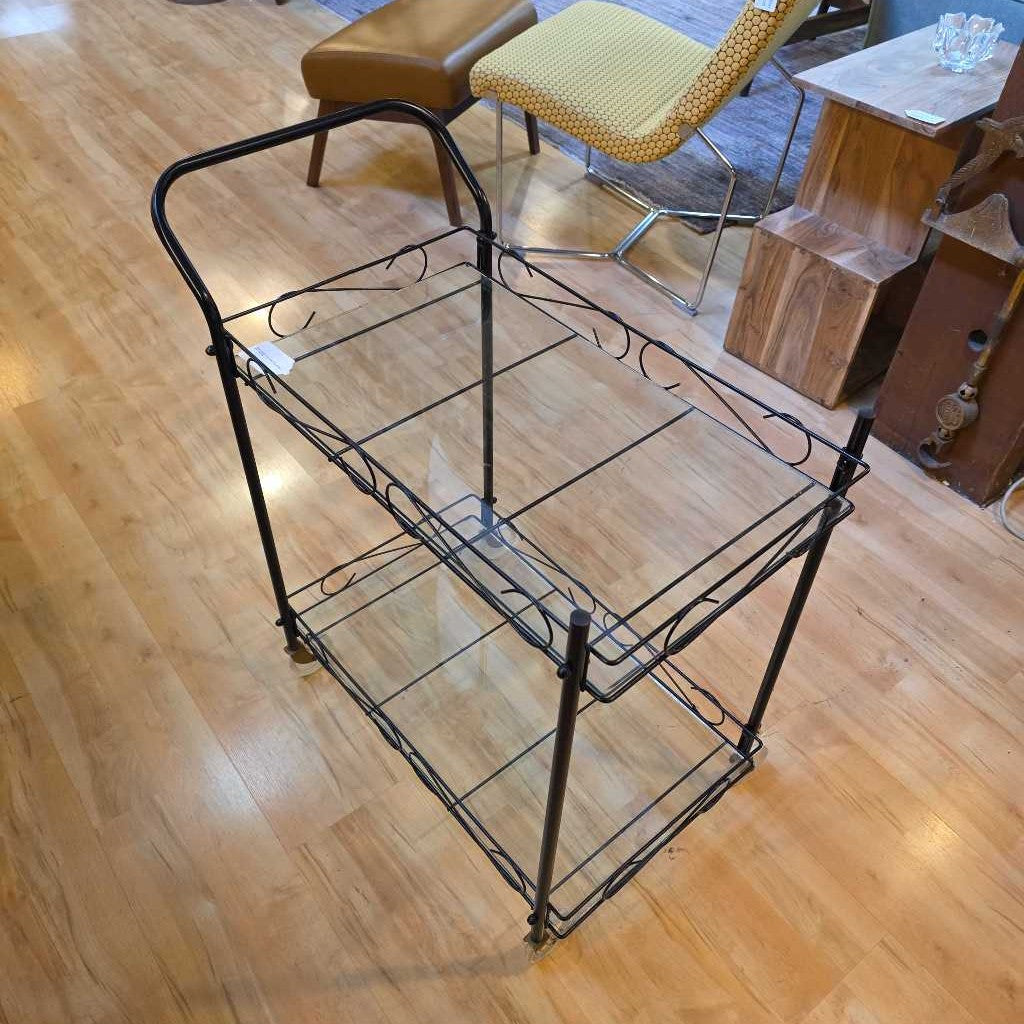 Black metal serving cart on a wooden floor with chairs and a table in the background.