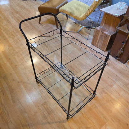 Black metal serving cart on a wooden floor with chairs and a table in the background.