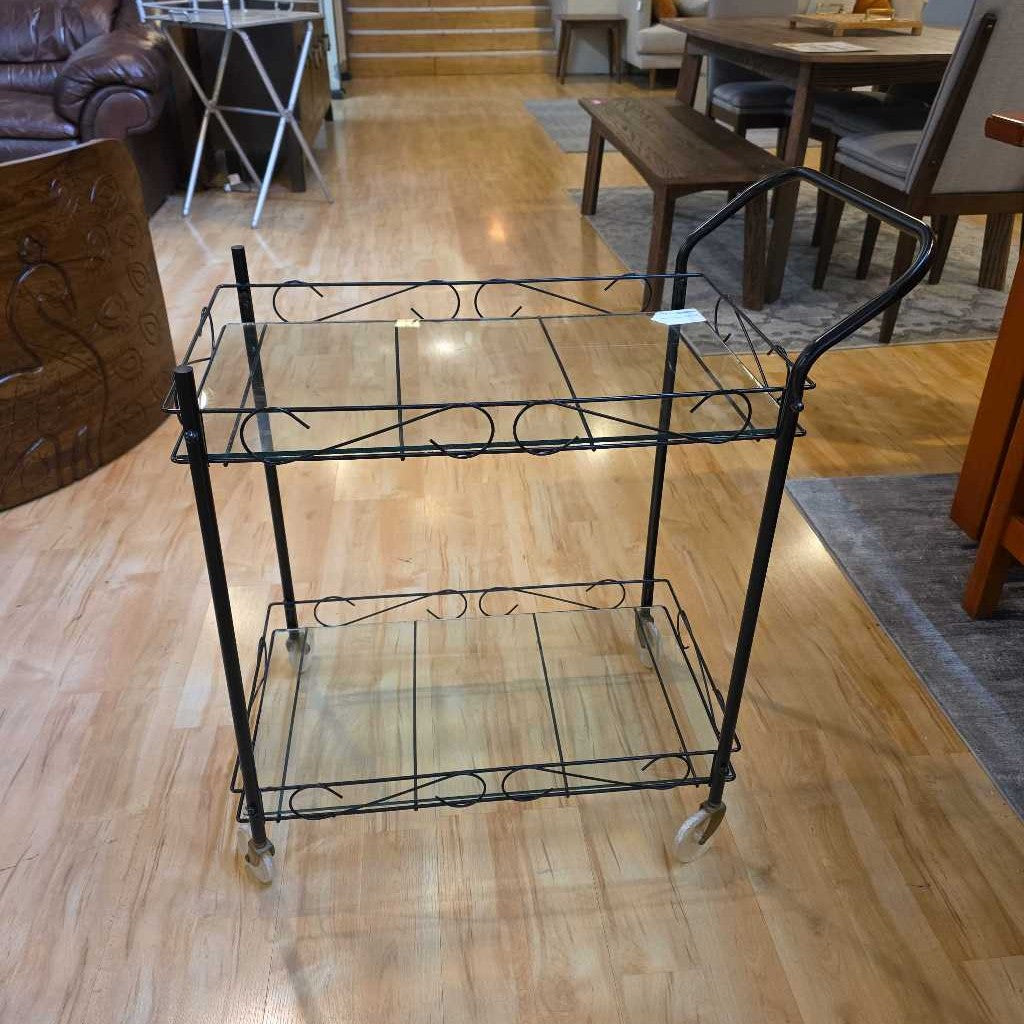 Two-tiered metal serving cart on wheels in a room with wooden flooring and furniture.