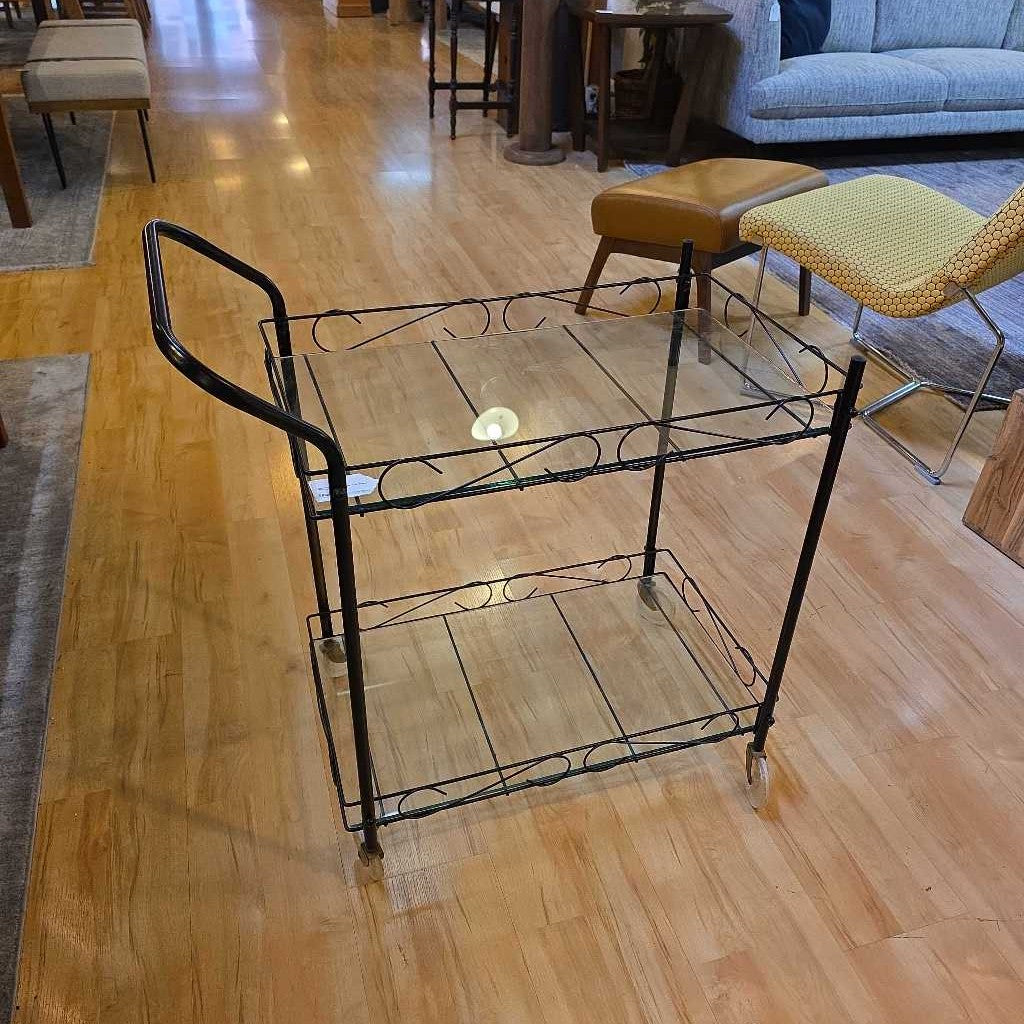 Wrought iron serving cart on a wooden floor with chairs and a couch in the background.