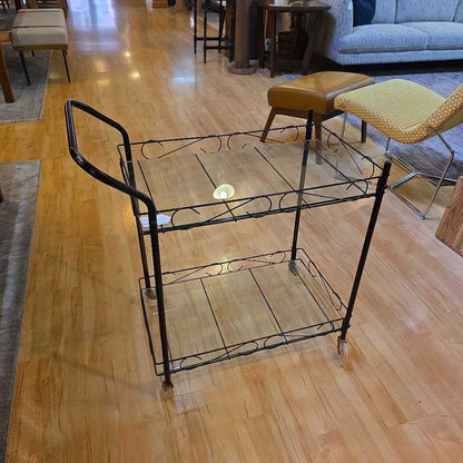 Wrought iron serving cart on a wooden floor with chairs and a couch in the background.