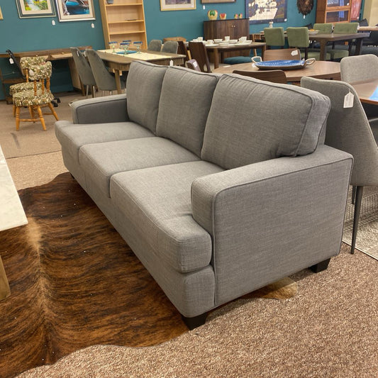 Gray sofa in a furniture store setting with various other furniture pieces in the background.