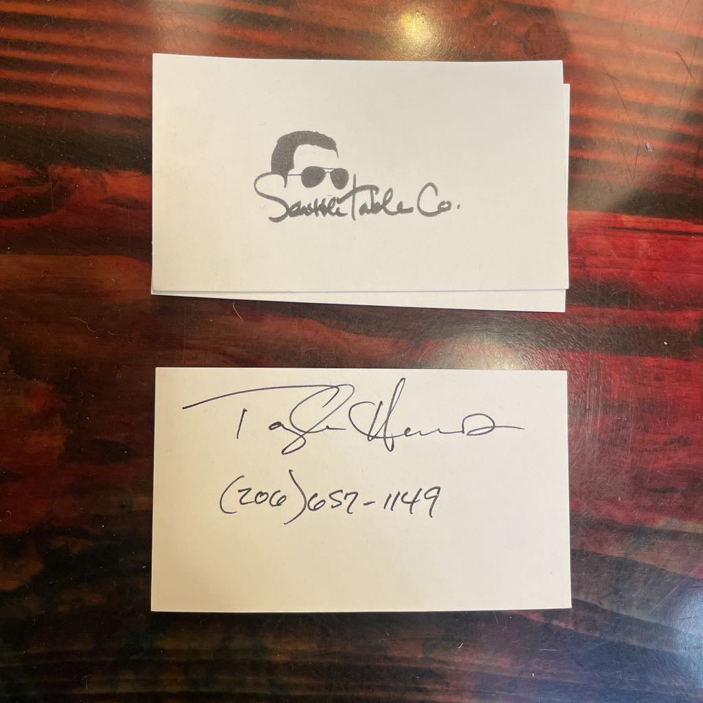 Two business cards on a wooden surface, one with a logo and text, the other with a signature.