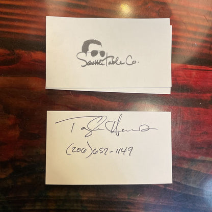Two business cards on a wooden surface, one with a logo and text, the other with a signature.