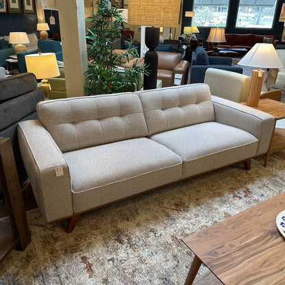 Beige sofa in a furniture store with various lamps and chairs in the background.