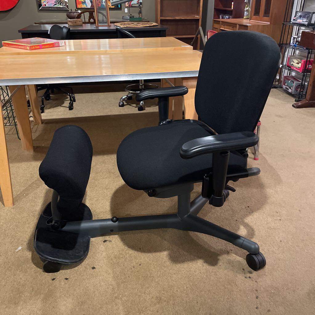 Black ergonomic chair in an office setting with a desk and chairs in the background.