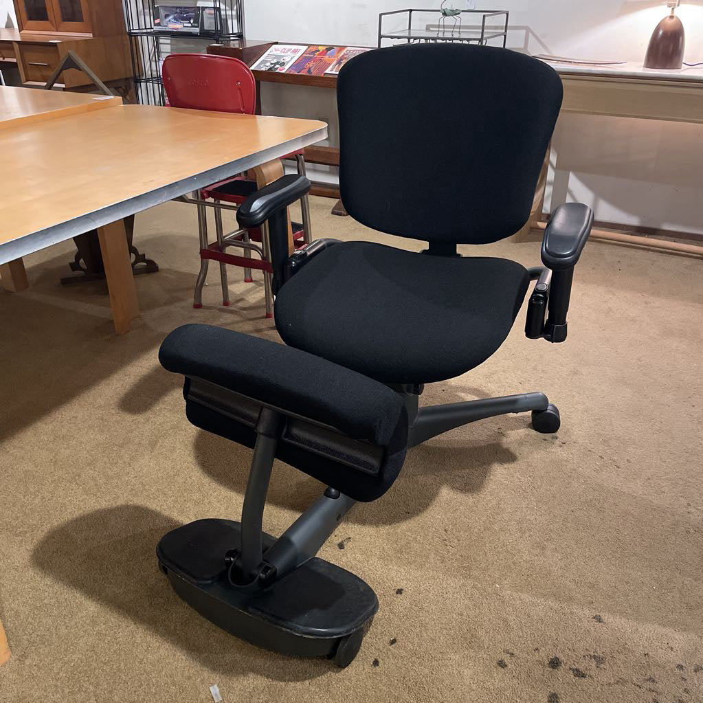 Black ergonomic chair on a beige carpeted floor with tables and chairs in the background.