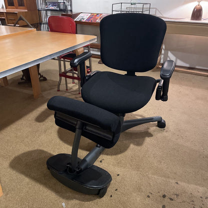 Black ergonomic chair on a beige carpeted floor with tables and chairs in the background.