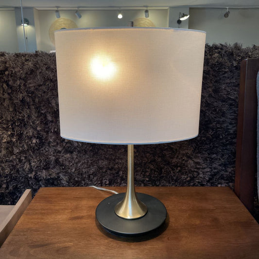 Table lamp with a beige shade on a wooden surface against a textured wall.