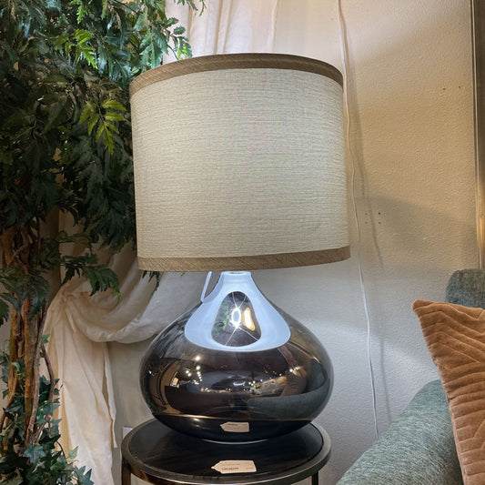 Table lamp with a beige shade on a black base next to a plant and draped fabric.