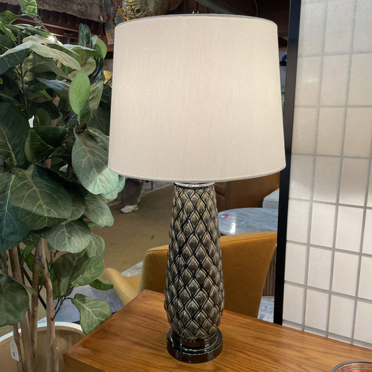 Decorative table lamp with textured base on a wooden surface next to a plant.