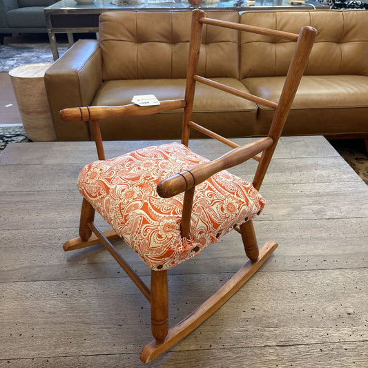 Wooden rocking chair with an orange paisley cushion on a wooden floor, with a brown sofa in the background.