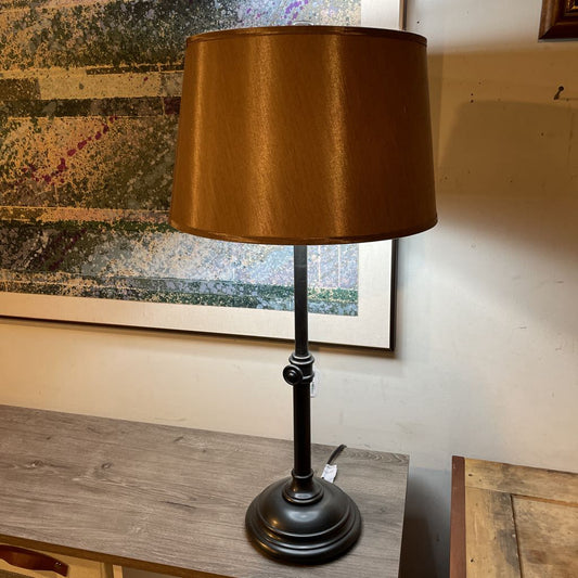Table lamp with a brown shade on a wooden surface, with a painting in the background.