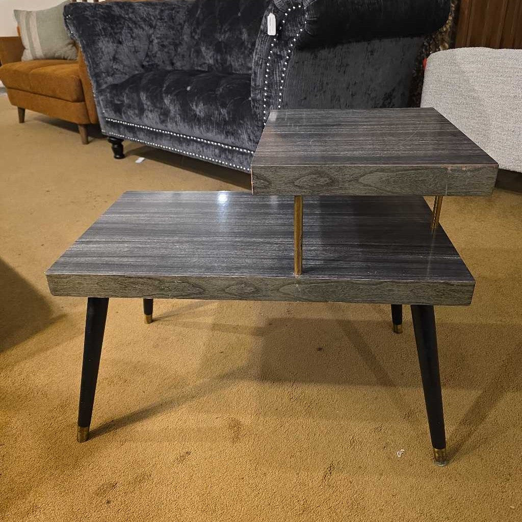 Two-tiered wooden table on a carpeted floor with a sofa in the background