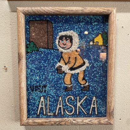 Framed artwork of a person in an Eskimo suit with 'Visit Alaska' text on a textured wall.
