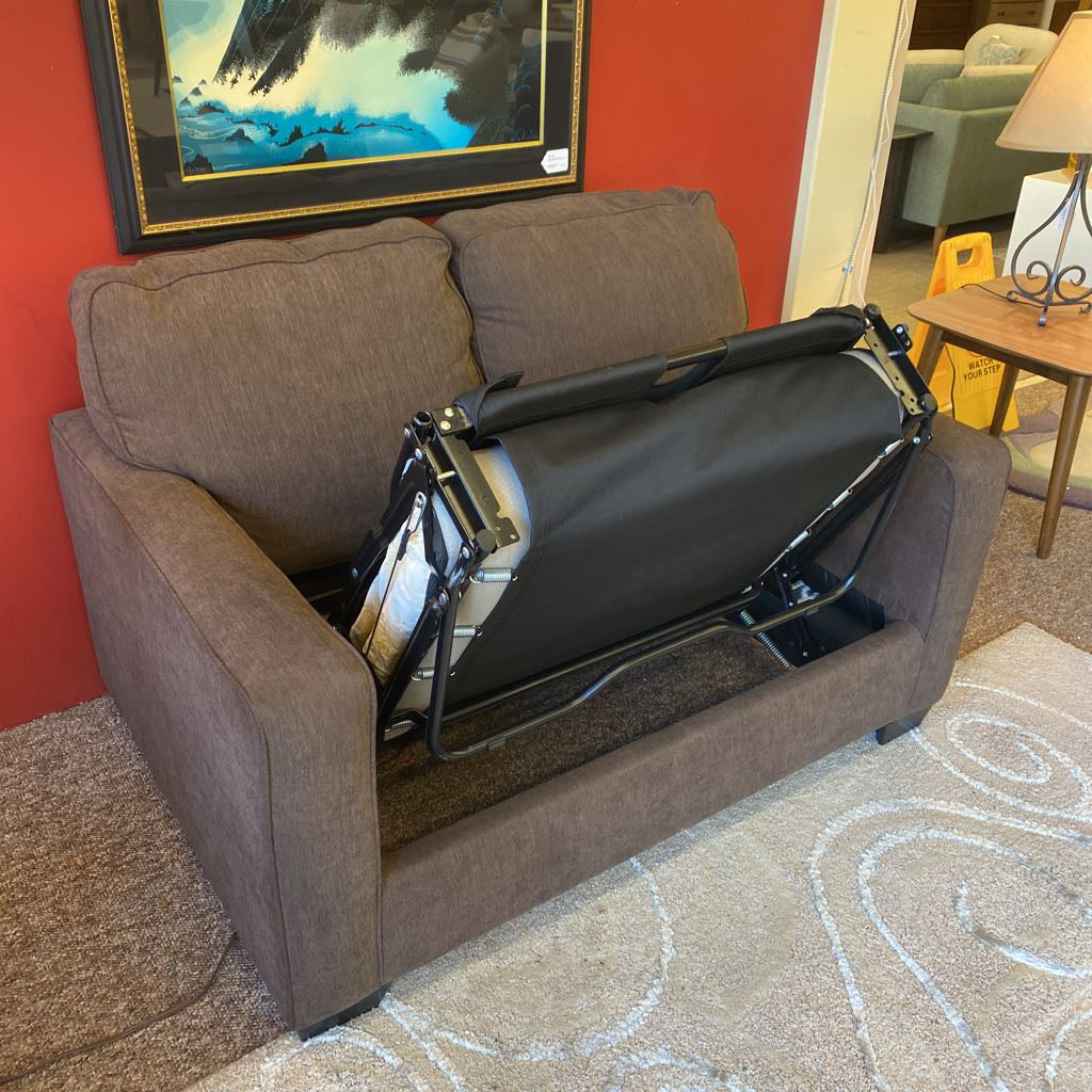 Brown sofa with a black office chair folded up on it in a room with a red wall and painting.