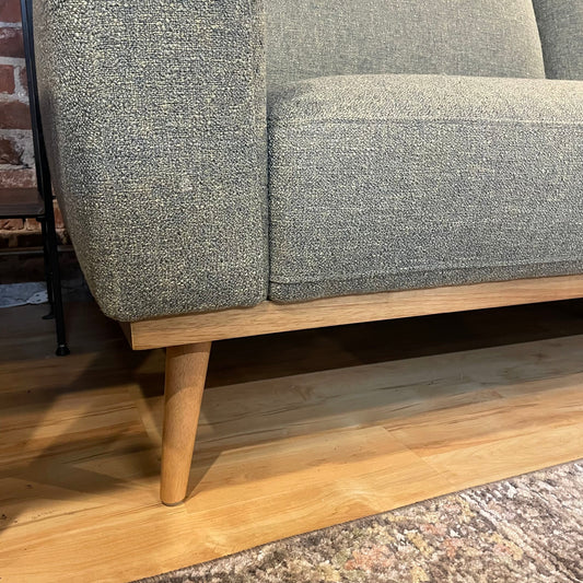 A light green textured sofa with two back cushions and two seat cushions, featuring a light wood base frame and tapered legs, set against an exposed red brick wall