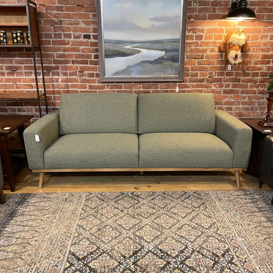 A light green textured sofa with two back cushions and two seat cushions, featuring a light wood base frame and tapered legs, set against an exposed red brick wall
