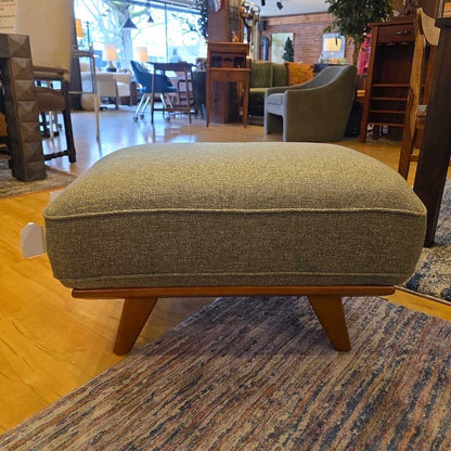 Gray upholstered ottoman with wooden legs in a furniture store setting