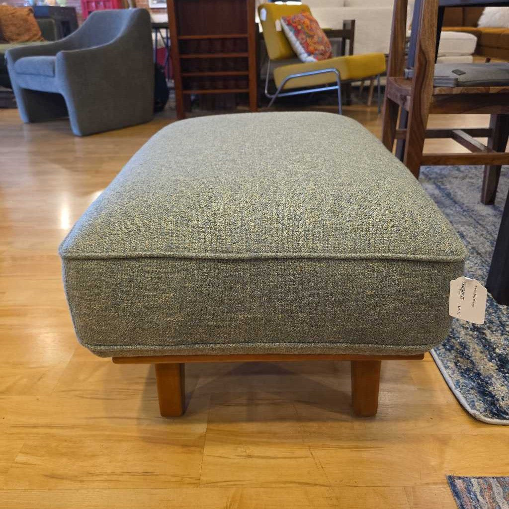 Green upholstered ottoman on a wooden floor with furniture in the background