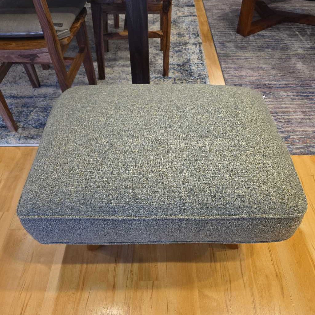Gray cushioned ottoman on a wooden floor with chairs and a rug in the background