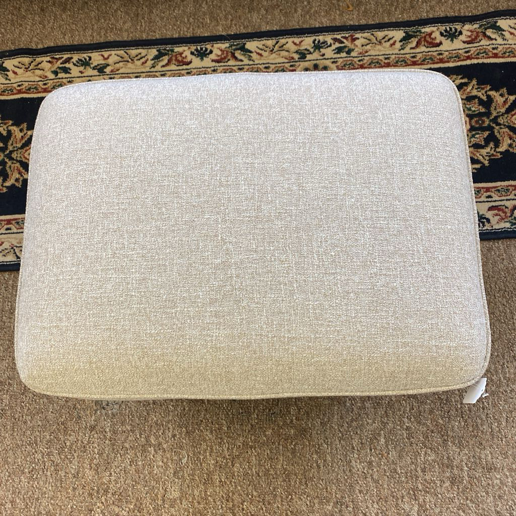 Beige ottoman on a carpeted floor with a decorative rug in the background
