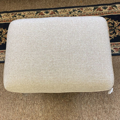 Beige ottoman on a carpeted floor with a decorative rug in the background