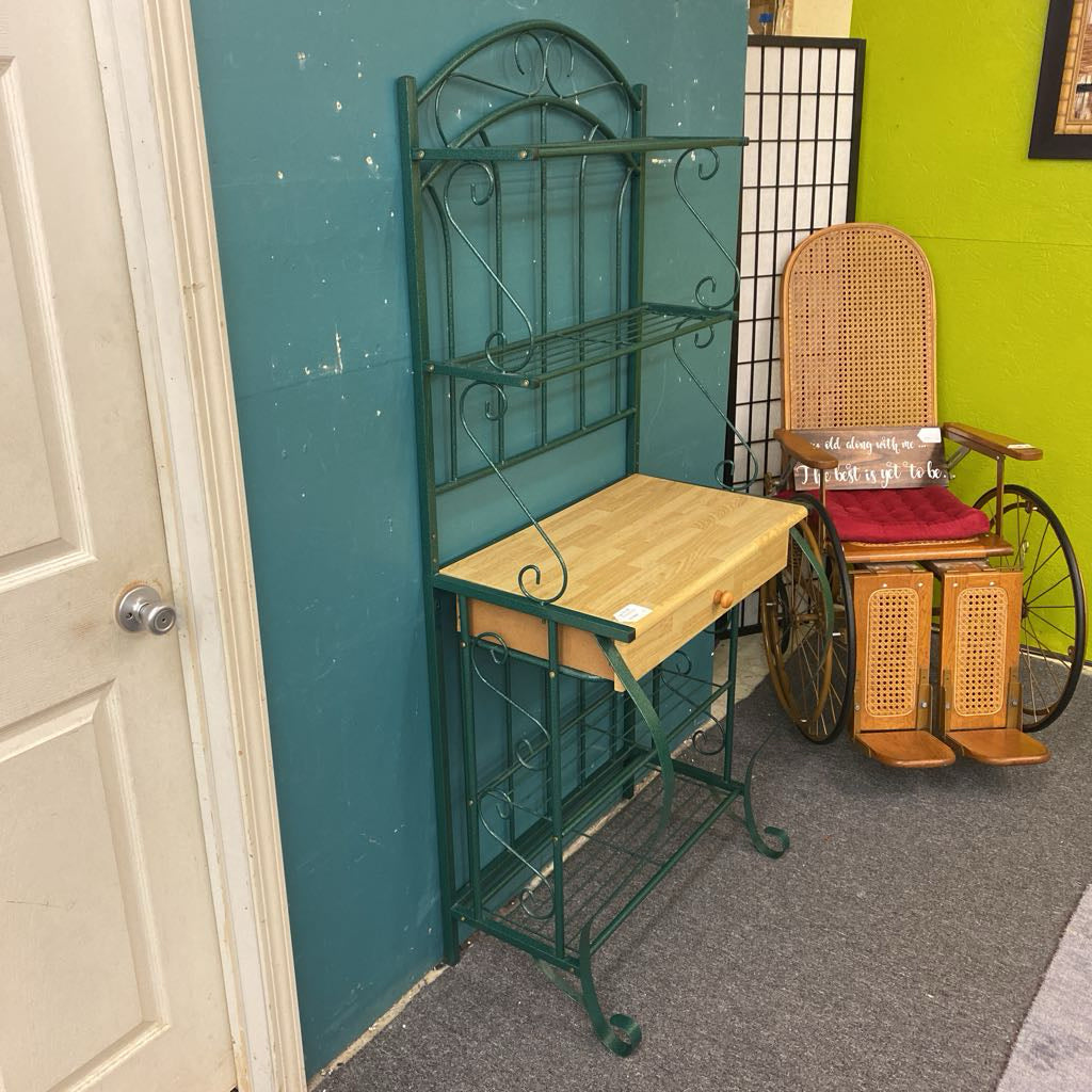 Green metal wall shelf with a wooden table against a teal wall, with a wicker chair and cart in the background.