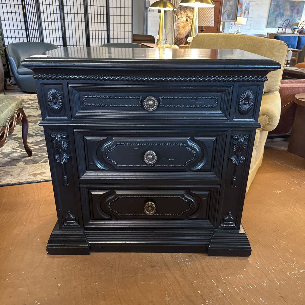 Decorative black nightstand with three drawers in a room setting.