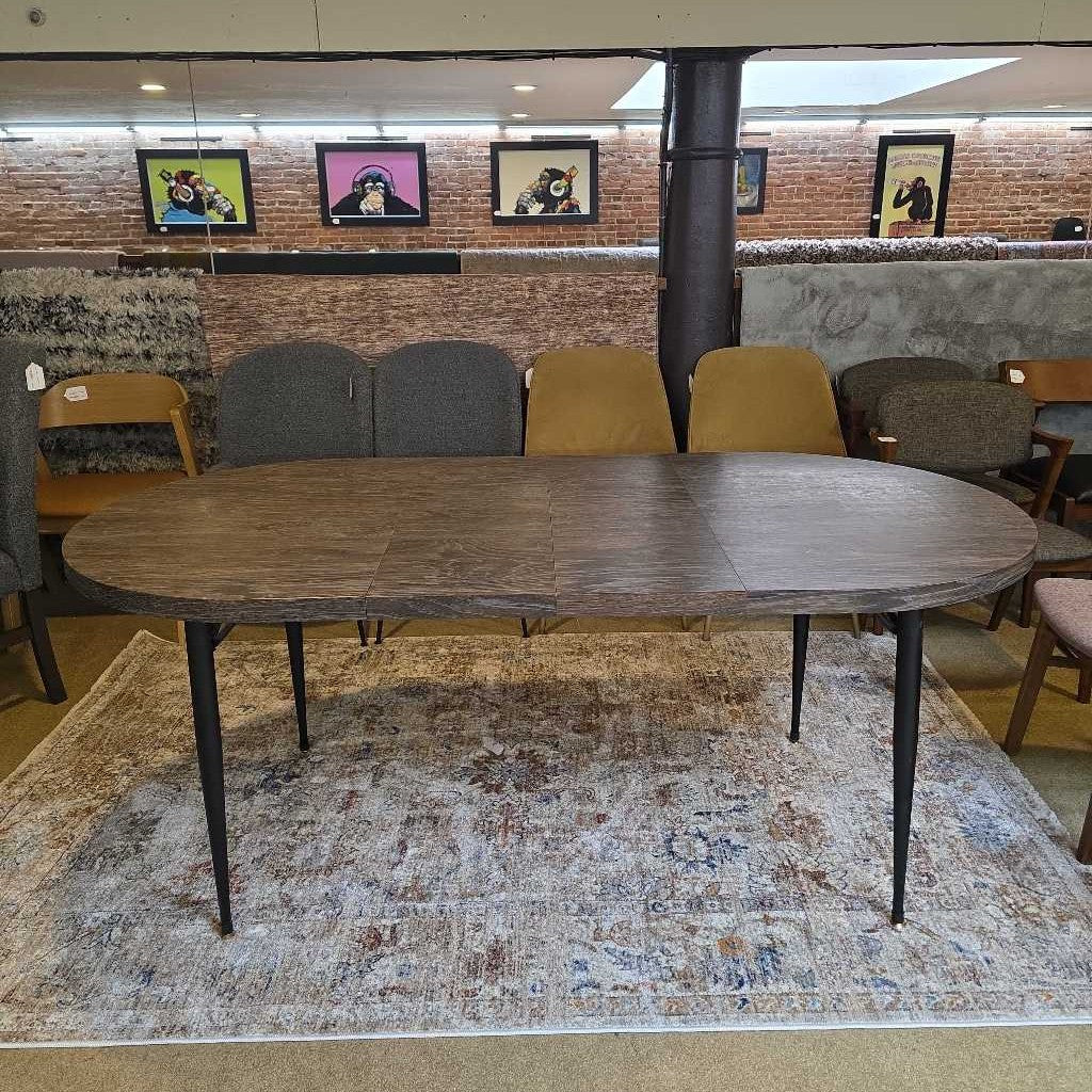 Wooden dining table with black legs on a patterned rug, surrounded by chairs in a room with framed pictures on the wall.