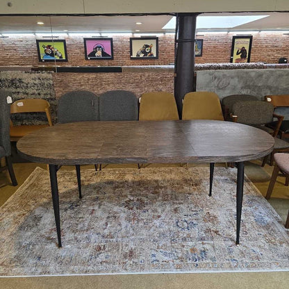 Wooden dining table with black legs on a patterned rug, surrounded by chairs in a room with framed pictures on the wall.