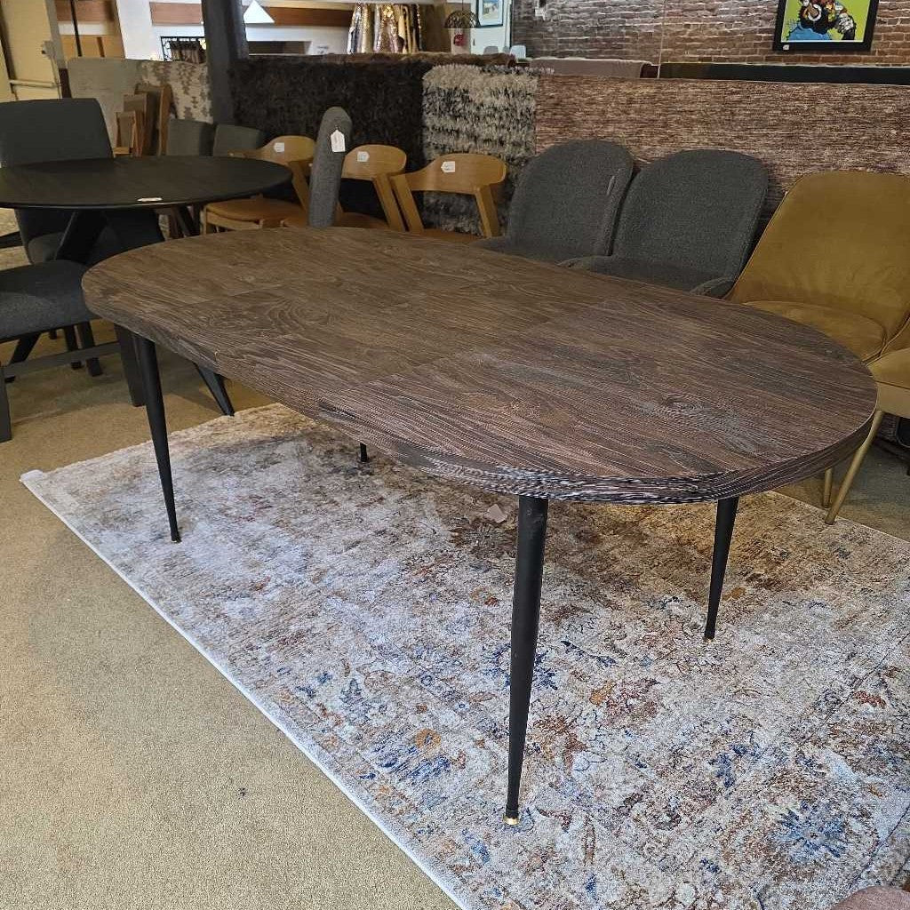 Wooden dining table with black legs on a patterned rug in a room setting.
