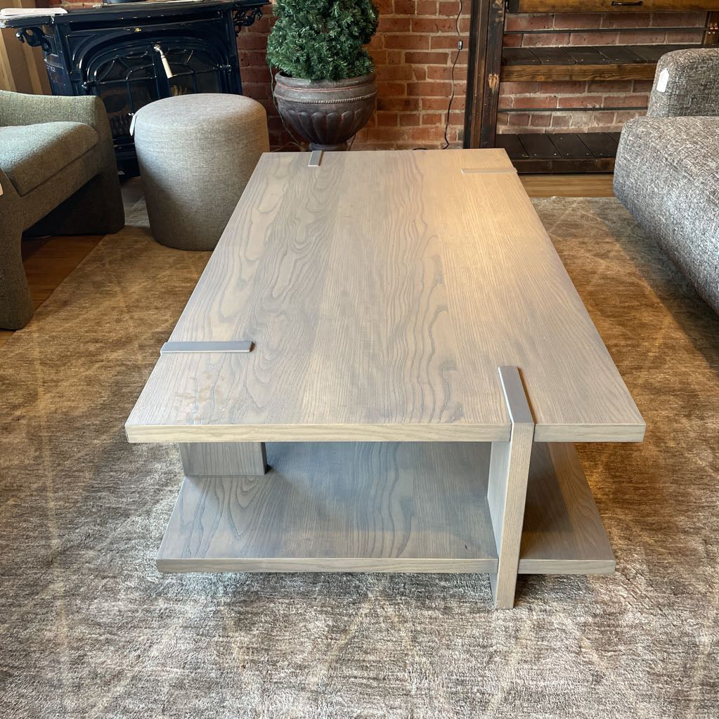 A modern rectangular coffee table with a grey wash wood finish, featuring a unique design where the support legs intersect and wrap over the edges of the top and bottom shelf