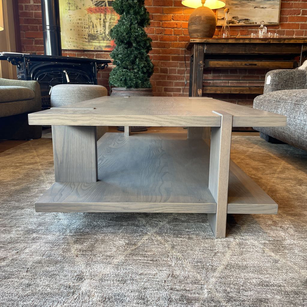 A modern rectangular coffee table with a grey wash wood finish, featuring a unique design where the support legs intersect and wrap over the edges of the top and bottom shelf