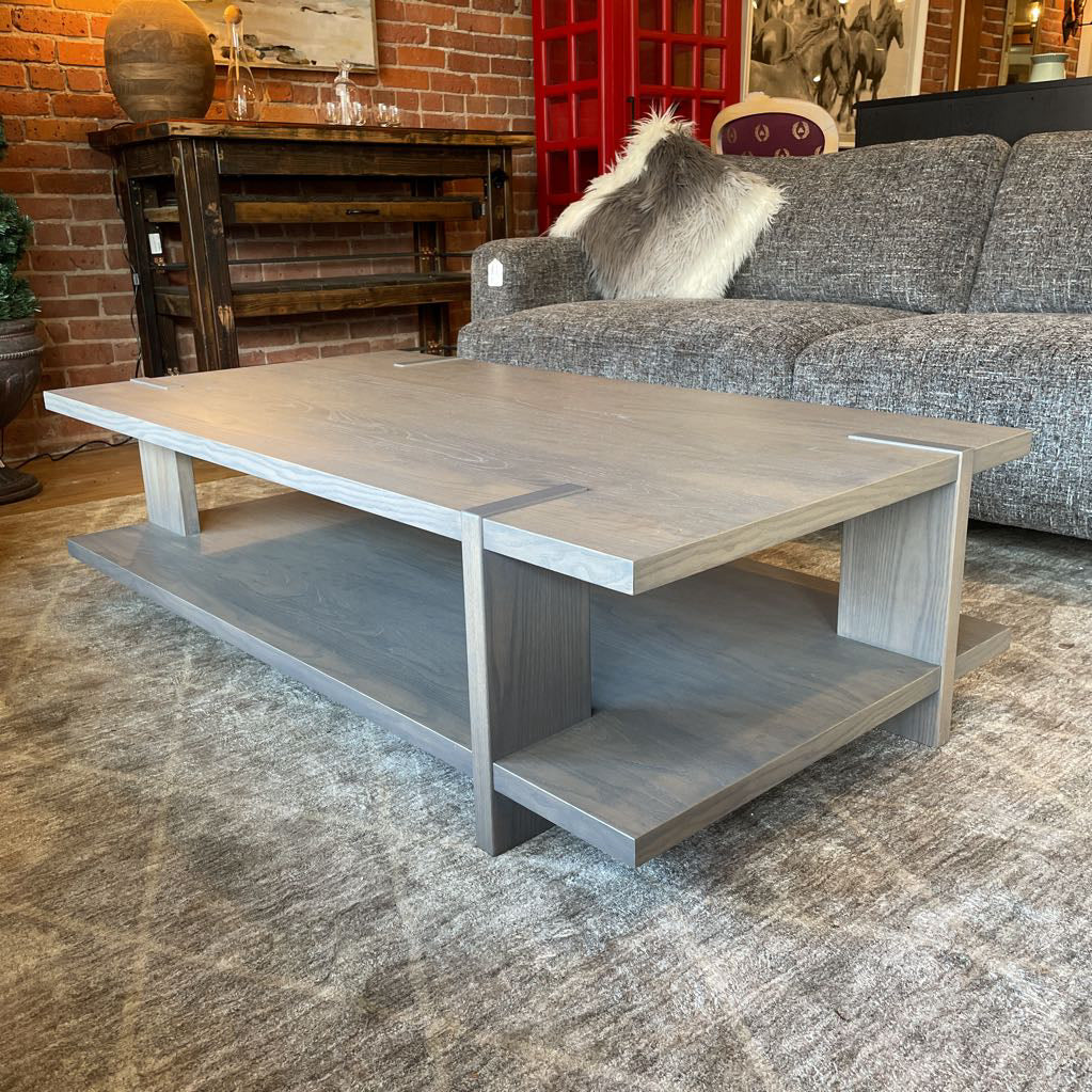 A modern rectangular coffee table with a grey wash wood finish, featuring a unique design where the support legs intersect and wrap over the edges of the top and bottom shelf
