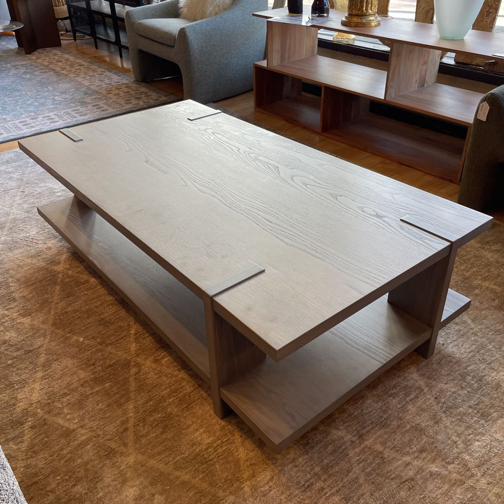 A modern rectangular coffee table with a grey wash wood finish, featuring a unique design where the support legs intersect and wrap over the edges of the top and bottom shelf