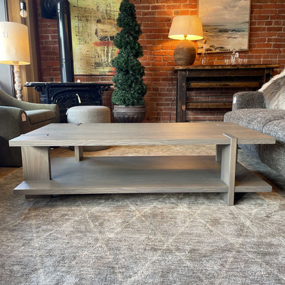 A modern rectangular coffee table with a grey wash wood finish, featuring a unique design where the support legs intersect and wrap over the edges of the top and bottom shelf