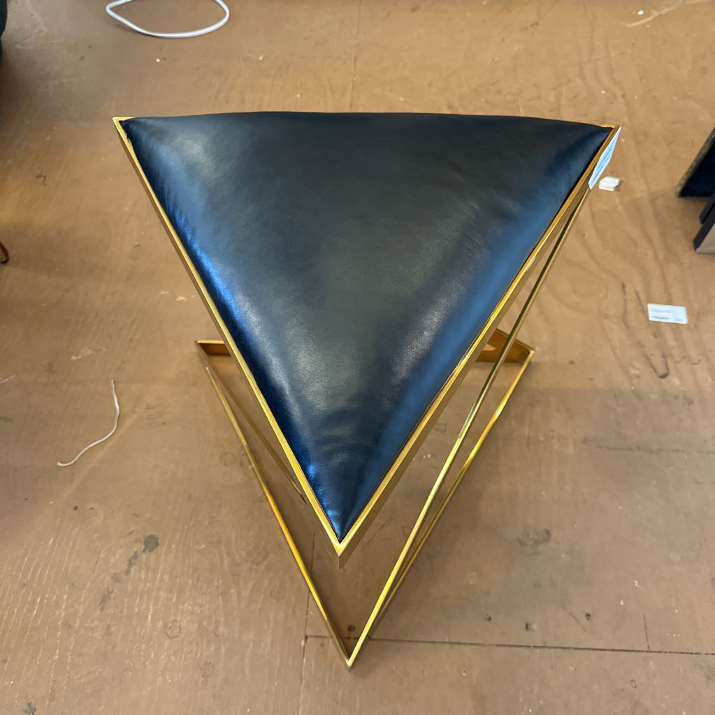 A modern geometric stool featuring a triangular black leather seat supported by a bold, X-shaped polished gold metal frame