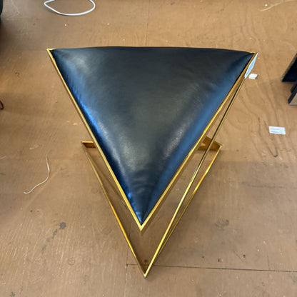A modern geometric stool featuring a triangular black leather seat supported by a bold, X-shaped polished gold metal frame
