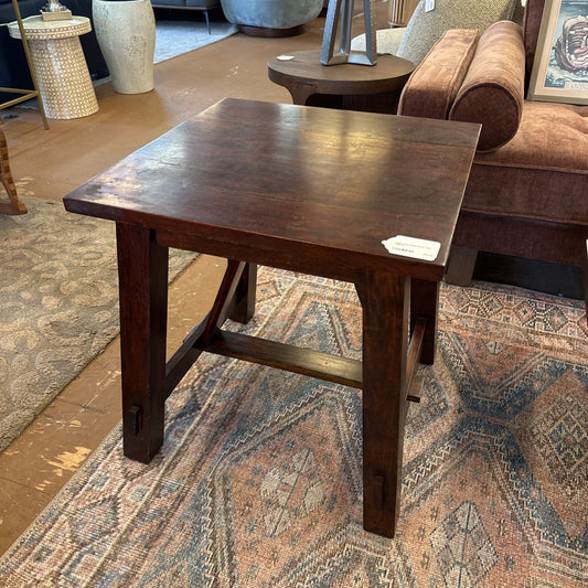 A square rustic side table with a dark, rich wood finish and substantial square legs joined by a low H-stretcher, resting on a patterned blue and cream area rug