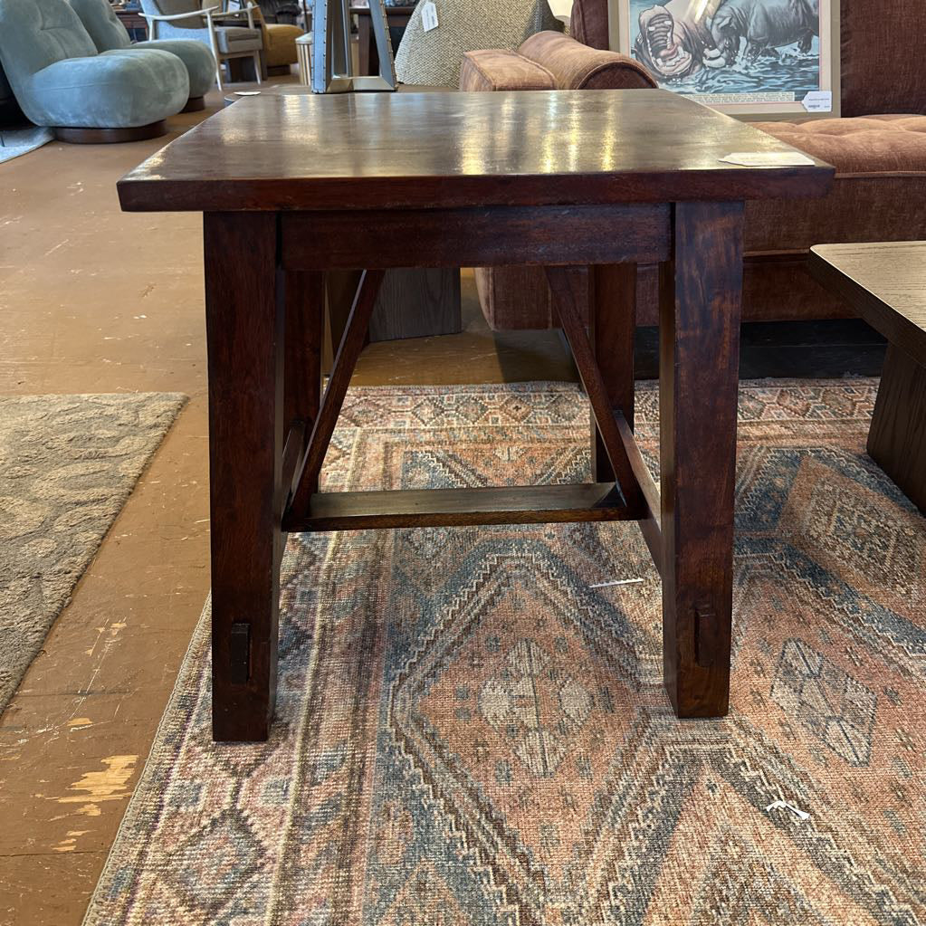 A square rustic side table with a dark, rich wood finish and substantial square legs joined by a low H-stretcher, resting on a patterned blue and cream area rug