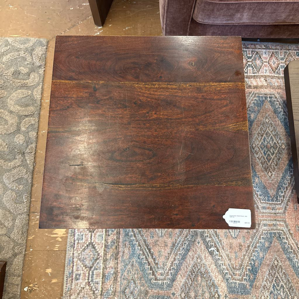 A square rustic side table with a dark, rich wood finish and substantial square legs joined by a low H-stretcher, resting on a patterned blue and cream area rug