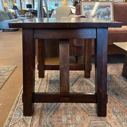 A square rustic side table with a dark, rich wood finish and substantial square legs joined by a low H-stretcher, resting on a patterned blue and cream area rug