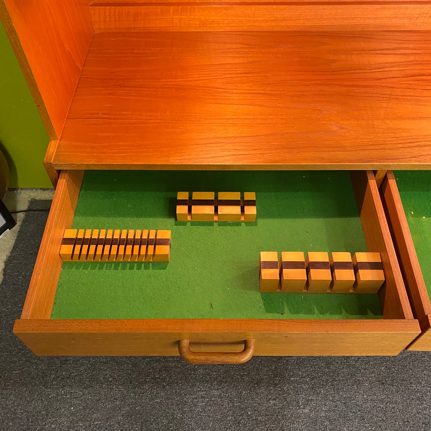Wooden drawer with green interior containing wooden blocks on a carpeted floor.