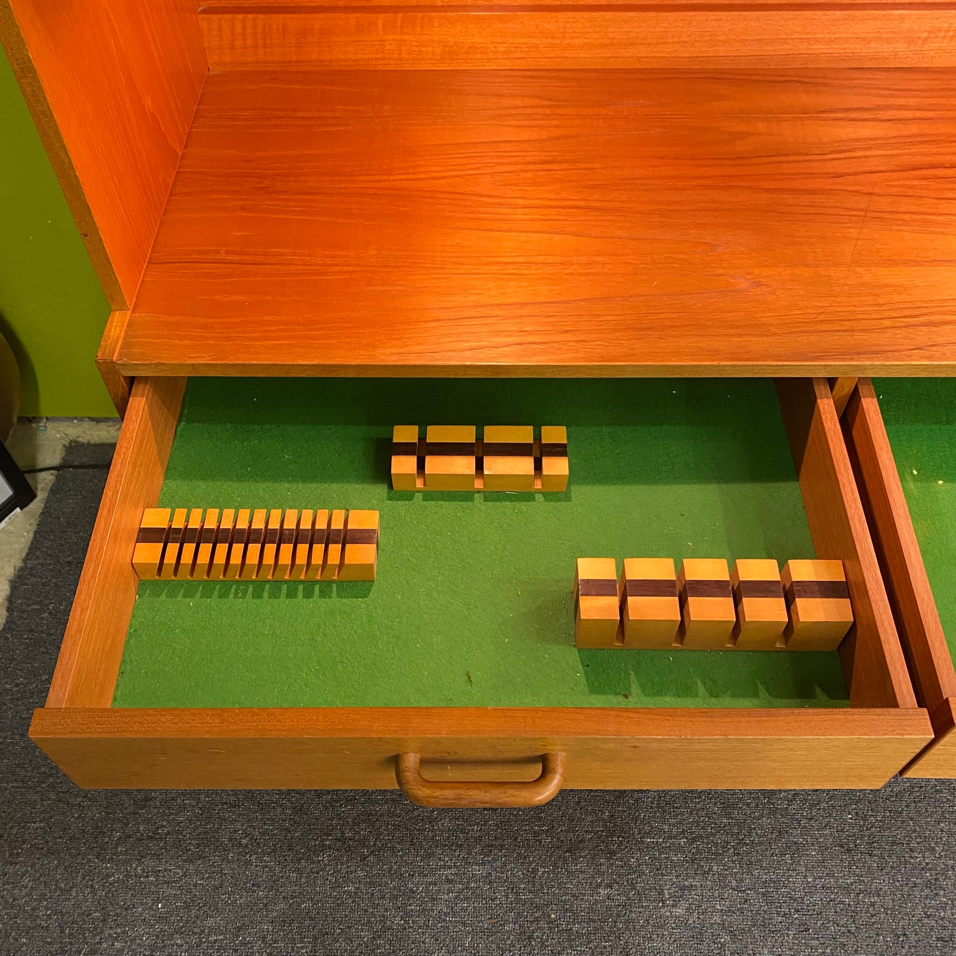 Wooden drawer with green interior containing wooden blocks on a carpeted floor.